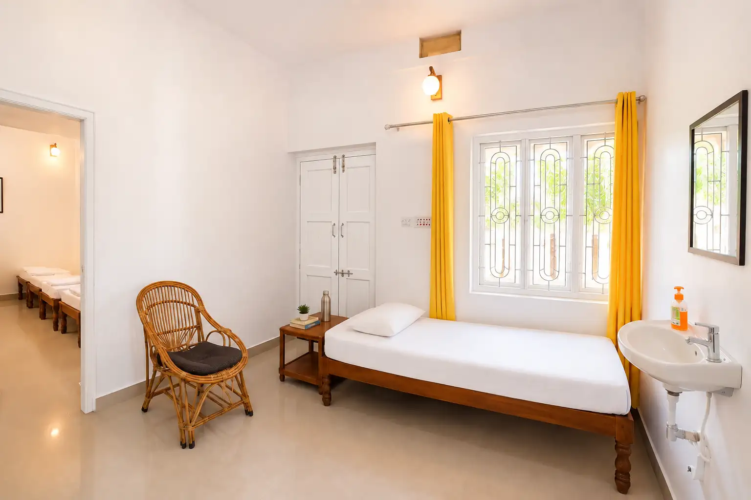 Wide view of single bed, cane chair, washbasin and doors in Nidhi sit-in room.